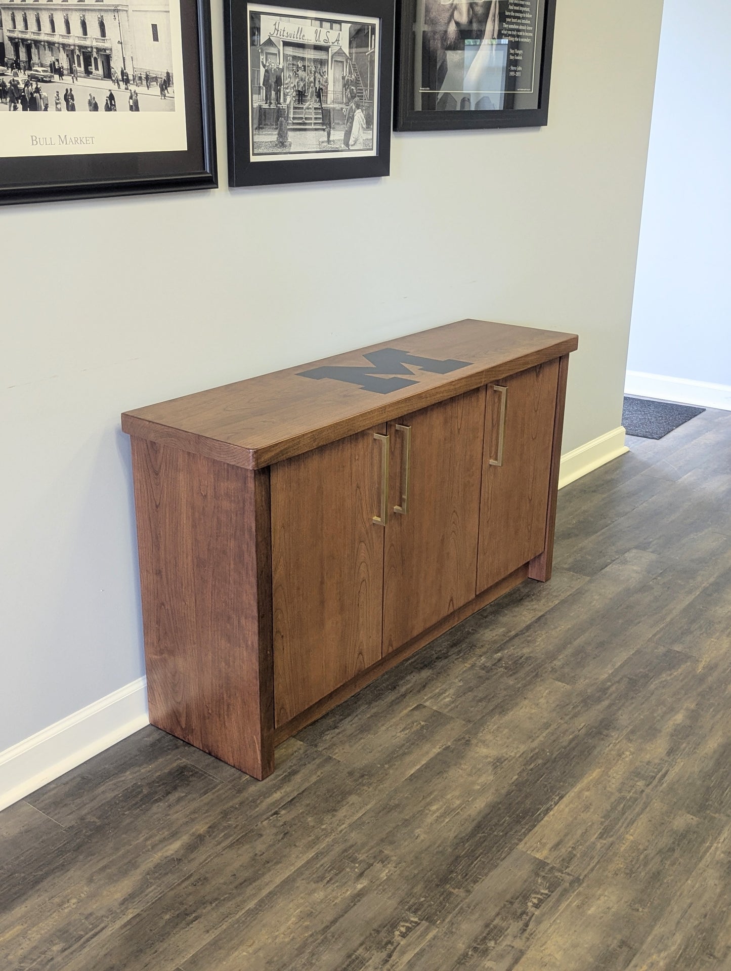 A modern stained cherry hardwood credenza with University of Michigan epoxy inlay on tabletop, three cabinets with gold handle finishes and interior storage for organizing office supplies.