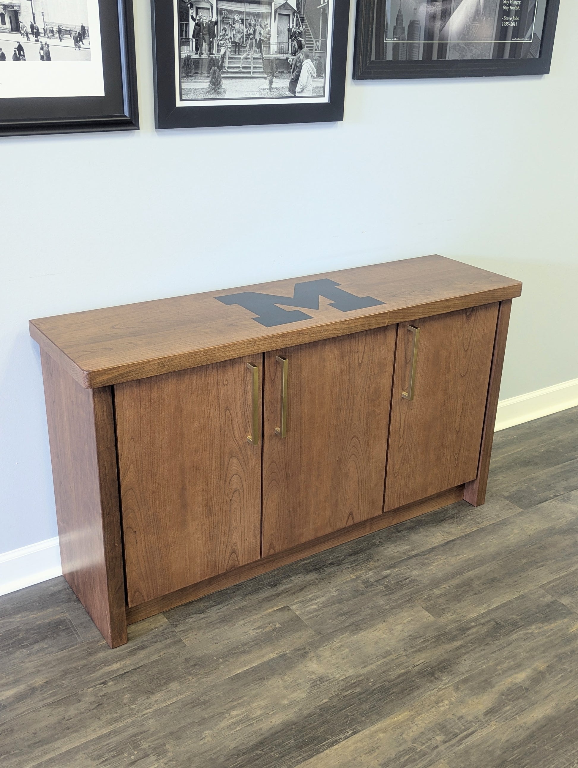 A modern stained cherry hardwood credenza with University of Michigan epoxy inlay on tabletop, three cabinets with gold handle finishes and interior storage for organizing office supplies.
