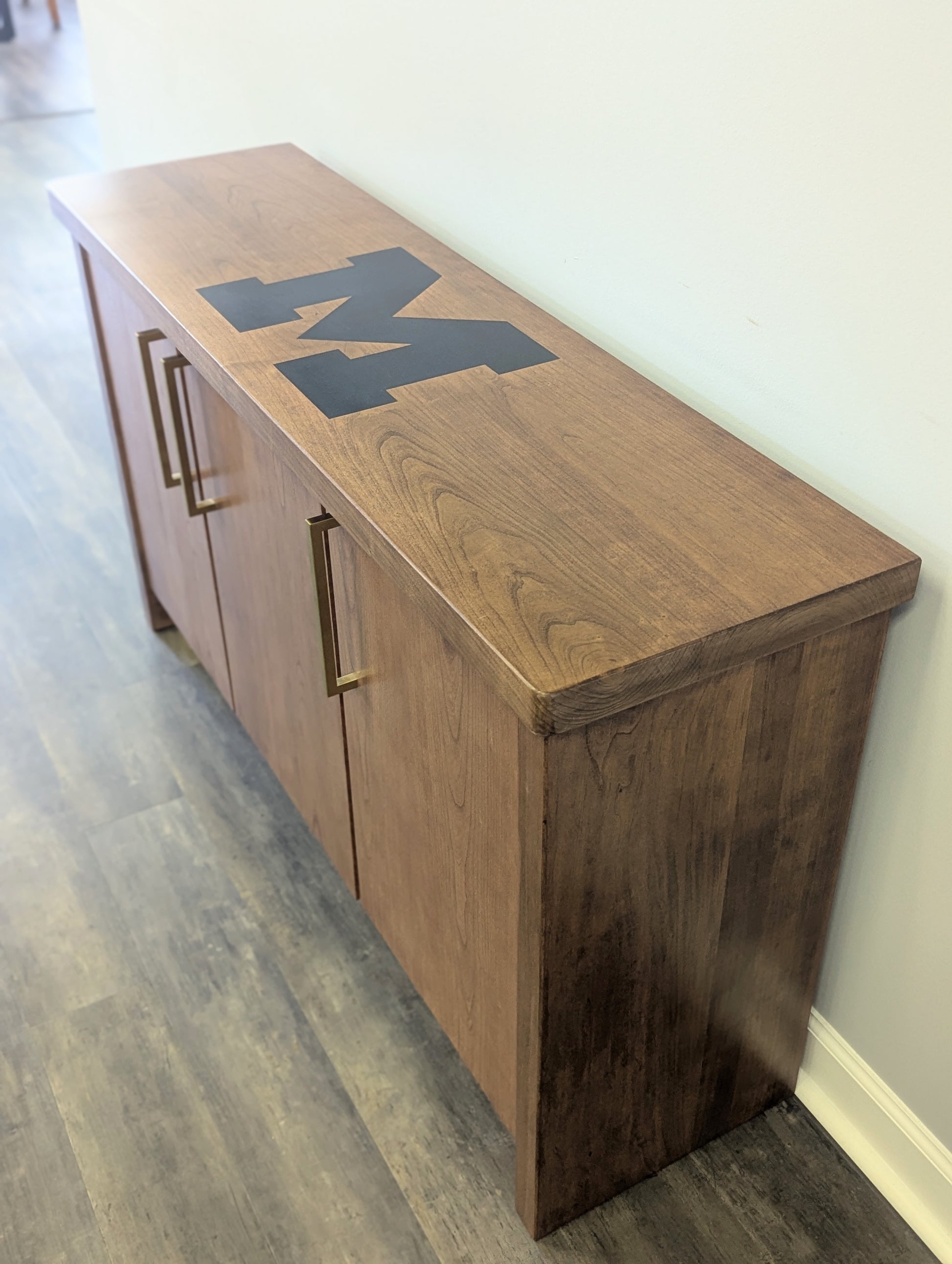 A modern stained cherry hardwood credenza with University of Michigan epoxy inlay on tabletop, three cabinets with gold handle finishes and interior storage for organizing office supplies.