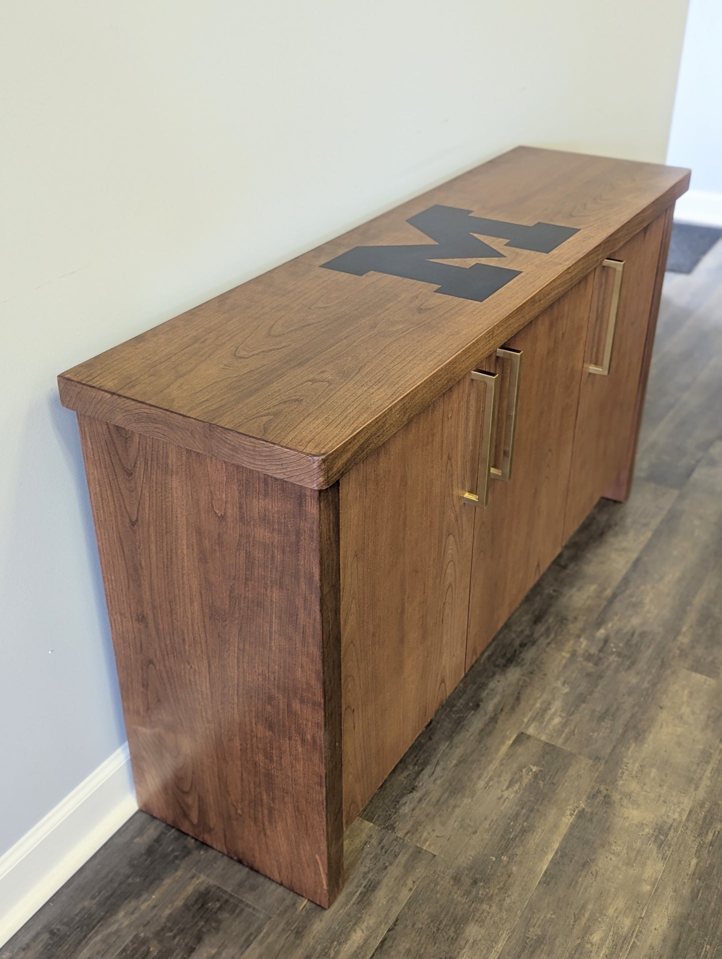 A modern stained cherry hardwood credenza with University of Michigan epoxy inlay on tabletop, three cabinets with gold handle finishes and interior storage for organizing office supplies.