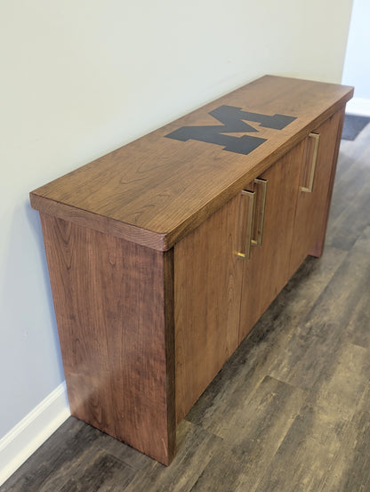 A modern stained cherry hardwood credenza with University of Michigan epoxy inlay on tabletop, three cabinets with gold handle finishes and interior storage for organizing office supplies.