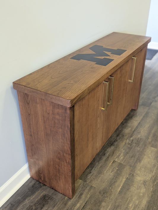 A modern stained cherry hardwood credenza with University of Michigan epoxy inlay on tabletop, three cabinets with gold handle finishes and interior storage for organizing office supplies.