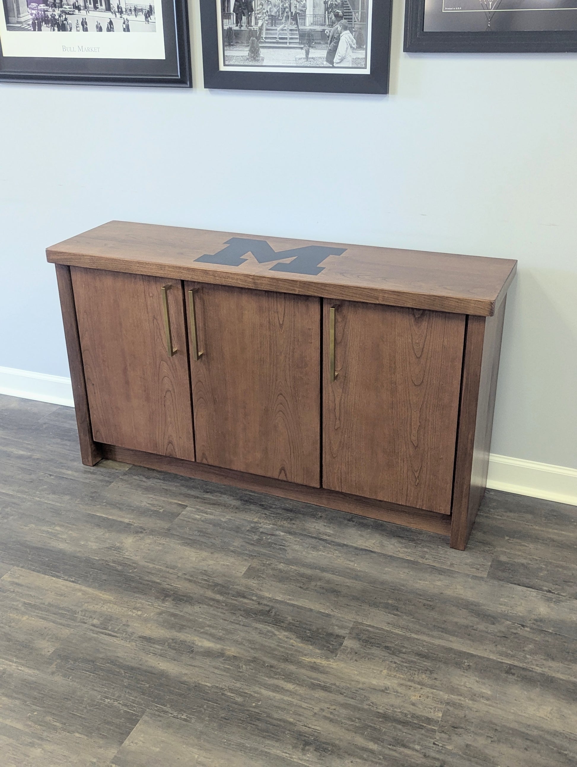 A modern stained cherry hardwood credenza with University of Michigan epoxy inlay on tabletop, three cabinets with gold handle finishes and interior storage for organizing office supplies.