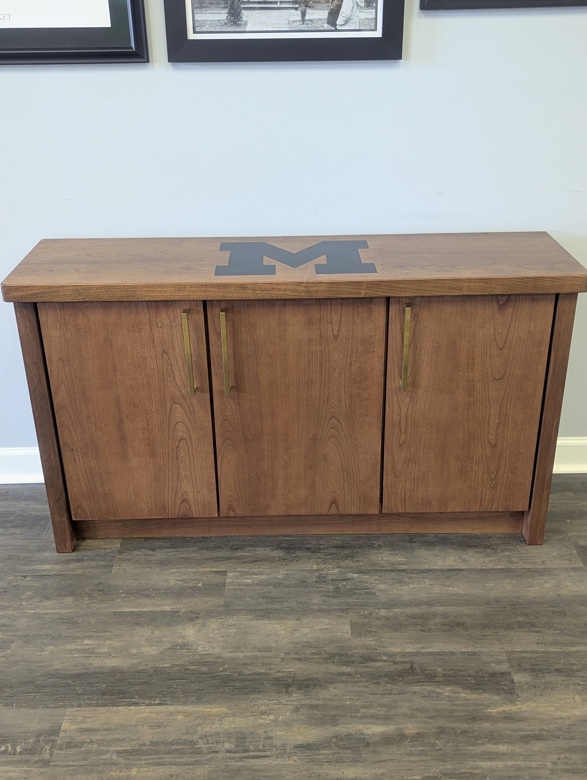 A modern stained cherry hardwood credenza with University of Michigan epoxy inlay on tabletop, three cabinets with gold handle finishes and interior storage for organizing office supplies.
