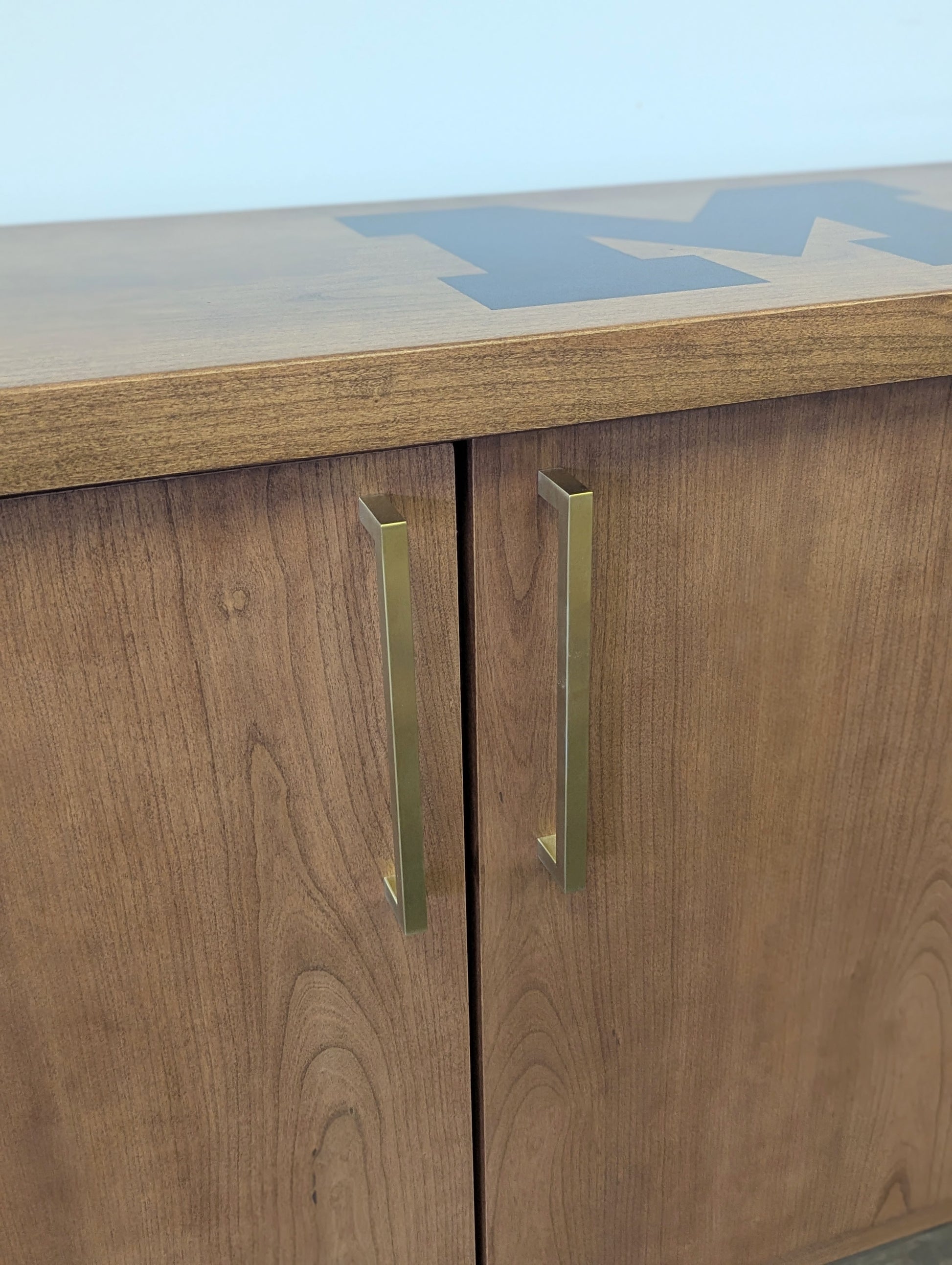A modern stained cherry hardwood credenza with University of Michigan epoxy inlay on tabletop, three cabinets with gold handle finishes and interior storage for organizing office supplies.
