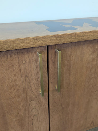 A modern stained cherry hardwood credenza with University of Michigan epoxy inlay on tabletop, three cabinets with gold handle finishes and interior storage for organizing office supplies.