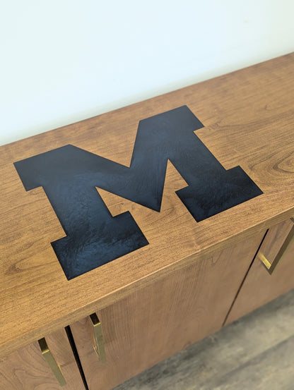 A modern stained cherry hardwood credenza with University of Michigan epoxy inlay on tabletop, three cabinets with gold handle finishes and interior storage for organizing office supplies.