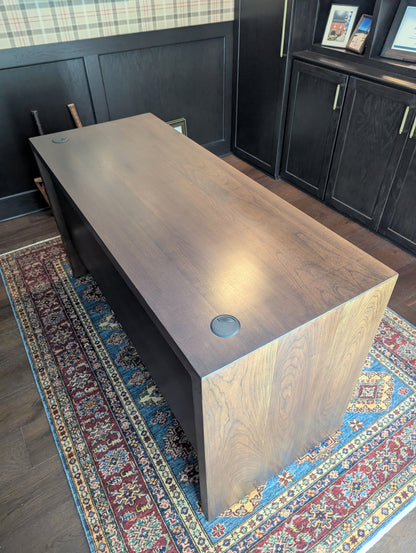 A modern stained cherry hardwood executive desk with waterfall style sides, grommets in the desk top and a hidden cord management shelf for clean storage.