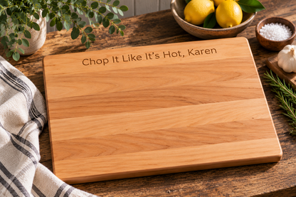A rectangular wooden cutting board with the engraved text 'Chop It Like It's Hot, Karen' on a rustic wooden table, surrounded by kitchen props including lemons, salt, garlic, rosemary, and a striped kitchen towel.