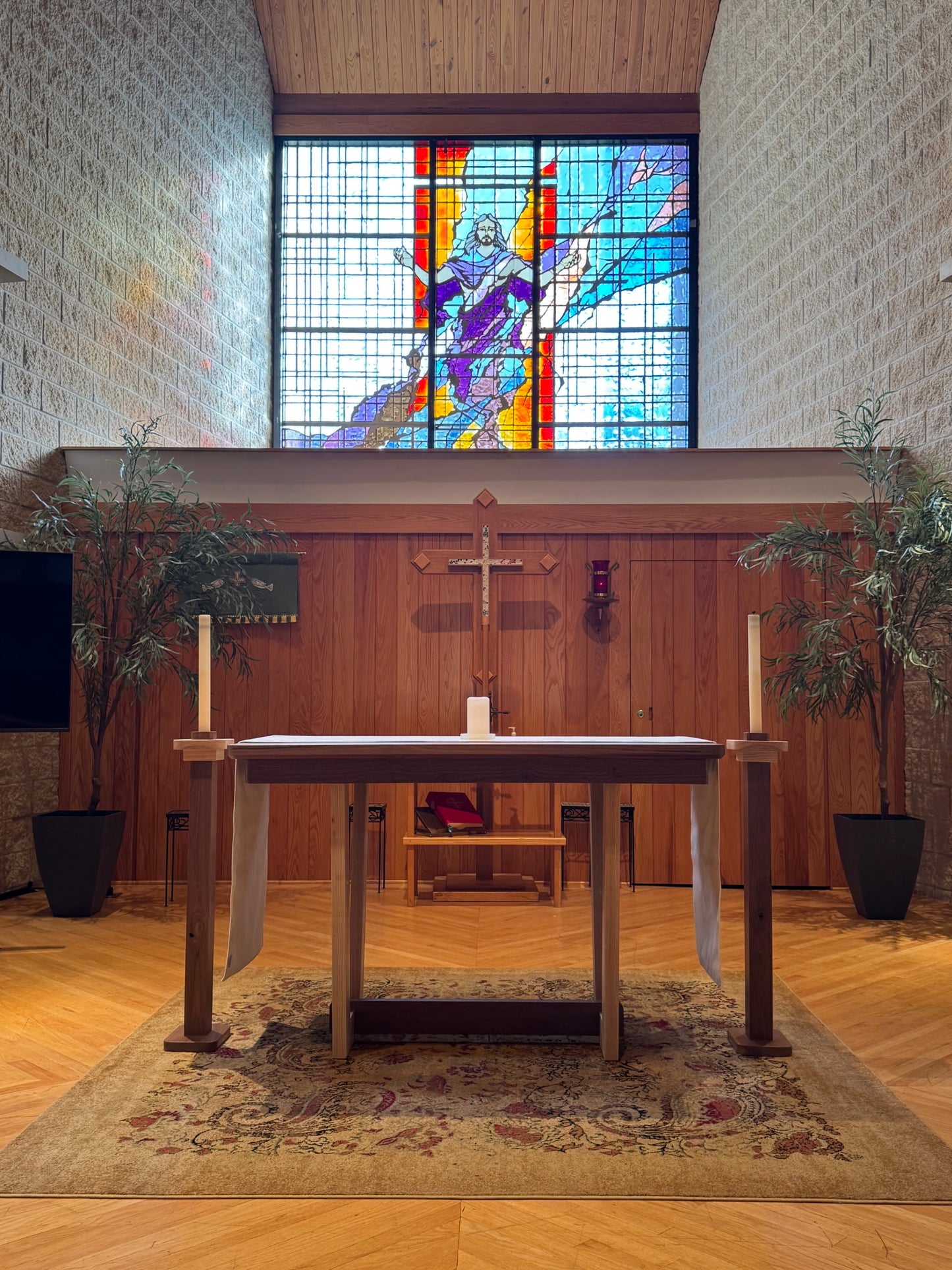 A wooden altar table with white draped cloths and tall white candles stands in a church-like interior with a stained-glass window showing a figure in purple and red robes, wood-paneled walls, and potted plants.