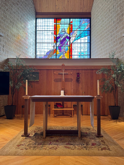 A wooden altar table with white draped cloths and tall white candles stands in a church-like interior with a stained-glass window showing a figure in purple and red robes, wood-paneled walls, and potted plants.