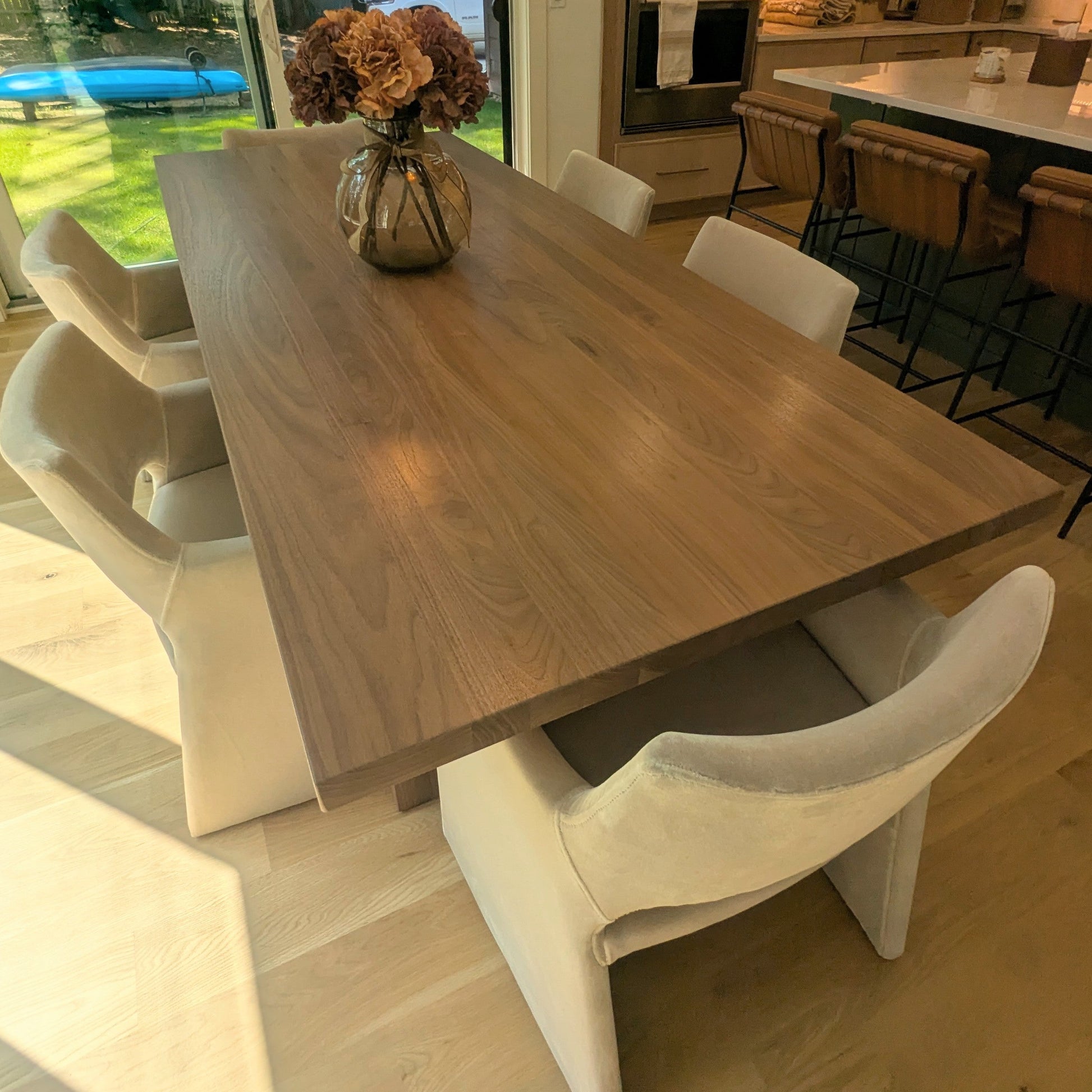 A rectangular wooden dining table with a warm medium-brown wood grain, surrounded by six modern off-white upholstered dining chairs. The table is styled with a small glass vase containing dried brown flowers, set in a bright, modern kitchen with light wood floors and a large window in the background.