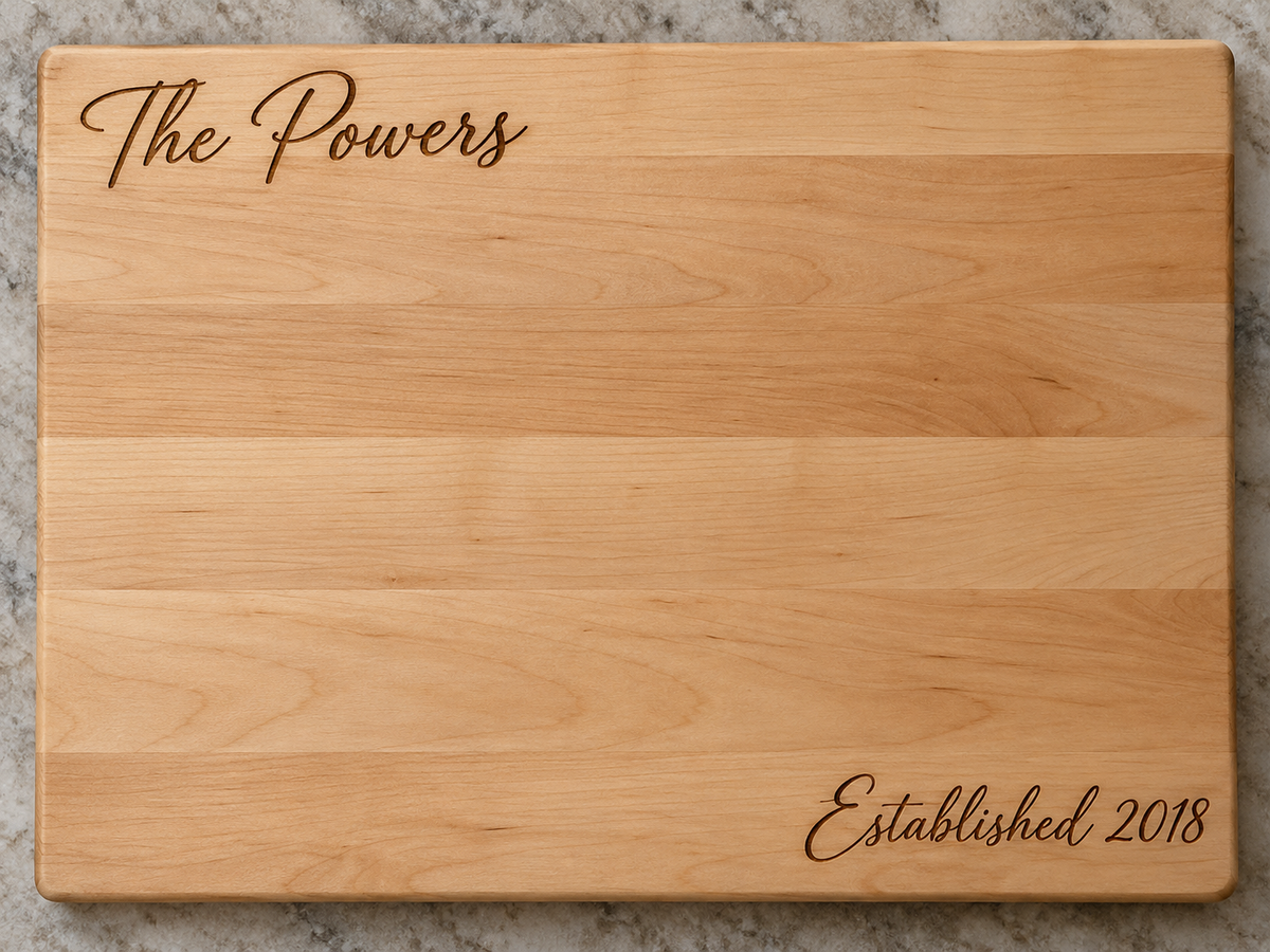 A rectangular wooden cutting board with a natural light wood grain finish, featuring the words 'The Powers' engraved in script at the top left and 'Established 2018' engraved in script at the bottom right, shown on a gray marble surface.