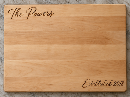 A rectangular wooden cutting board with a natural light wood grain finish, featuring the words 'The Powers' engraved in script at the top left and 'Established 2018' engraved in script at the bottom right, shown on a gray marble surface.