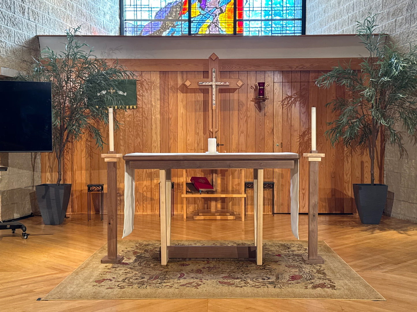 A wooden altar table with a draped cloth, set on a patterned rug inside a church or chapel with a wood-paneled wall, crucifix, stained glass window, and potted plants.