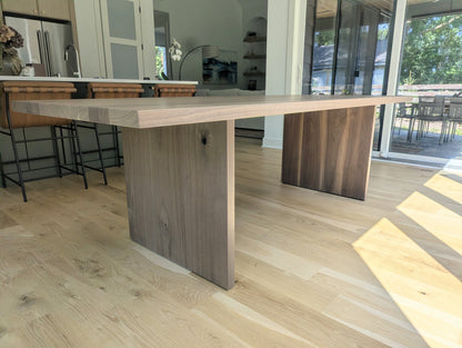 A modern rectangular dining table with a light natural wood top and thick block legs, shown in a bright contemporary kitchen with light wood floors and glass doors.