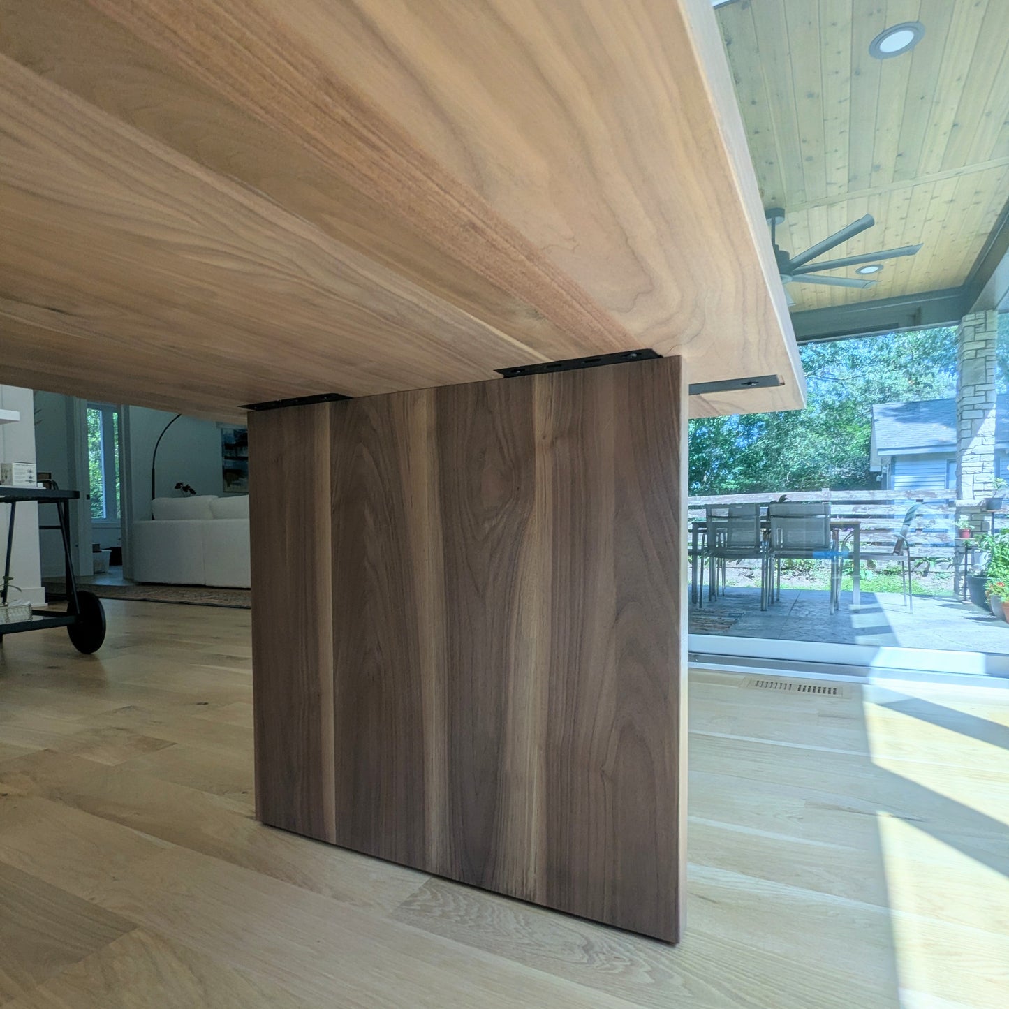 A close-up view of a wooden dining table base with a large light wood tabletop and a dark wood pedestal base, set in a modern home interior with light wood floors and a patio visible in the background.