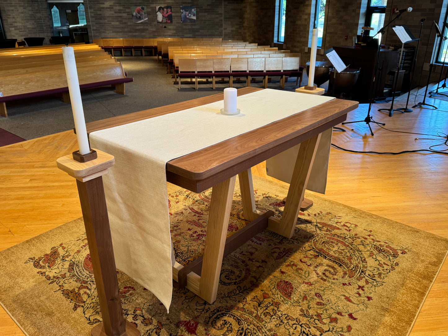 A wooden altar table with a white fabric runner and two white candles, set on a patterned rug inside a church hall with wooden pews and musical instruments in the background.