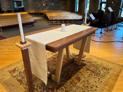 A wooden altar table with a white fabric runner and two white candles, set on a patterned rug inside a church hall with wooden pews and musical instruments in the background.