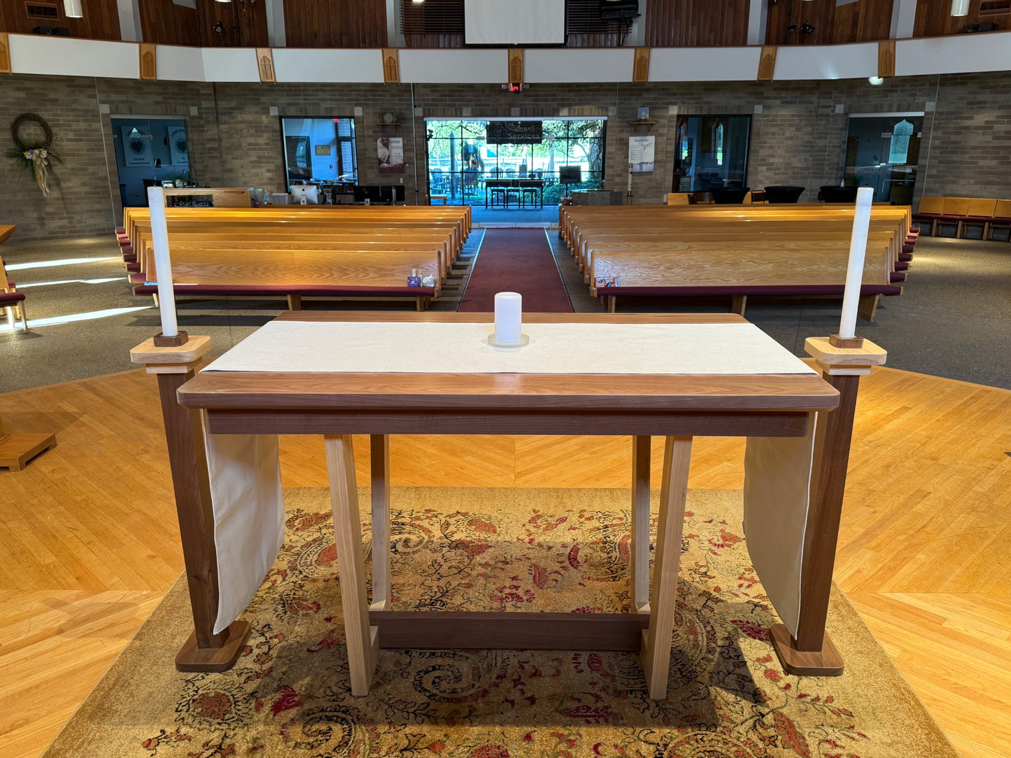 A wooden altar table with a white cloth top, draped side hangings, and two tall white candles on its ends, set in a church interior with pews, wood floors, and a patterned rug.