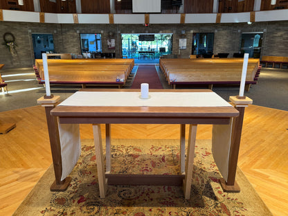 A wooden altar table with a white cloth top, draped side hangings, and two tall white candles on its ends, set in a church interior with pews, wood floors, and a patterned rug.