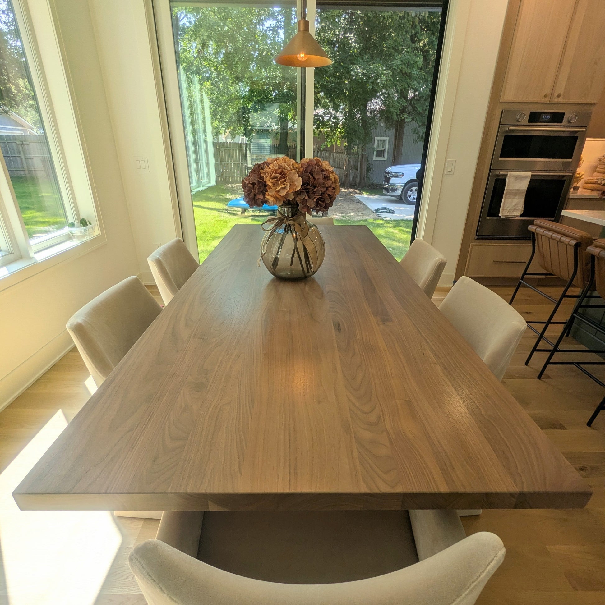 A long rectangular wooden dining table with a natural walnut finish, surrounded by light beige upholstered chairs in a bright modern dining room with large windows and pendant lights.