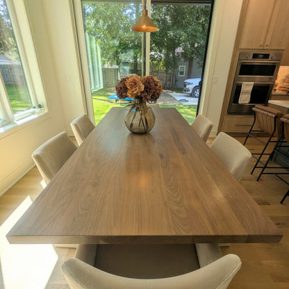 A long rectangular wooden dining table with a natural walnut finish, surrounded by light beige upholstered chairs in a bright modern dining room with large windows and pendant lights.
