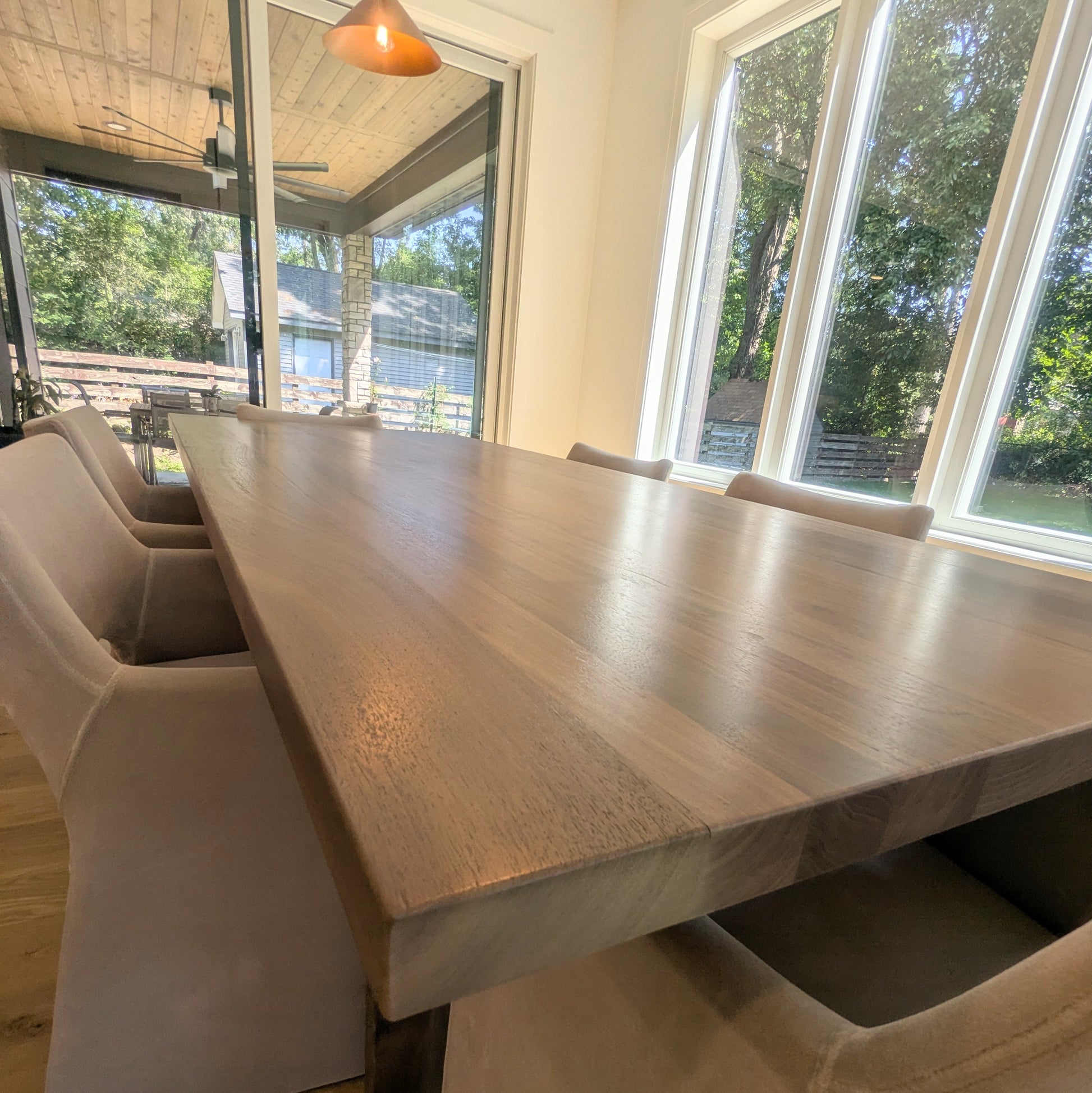 A long rectangular wooden dining table with a warm medium-brown finish, surrounded by upholstered dining chairs in a bright dining room with large windows and a visible outdoor patio.