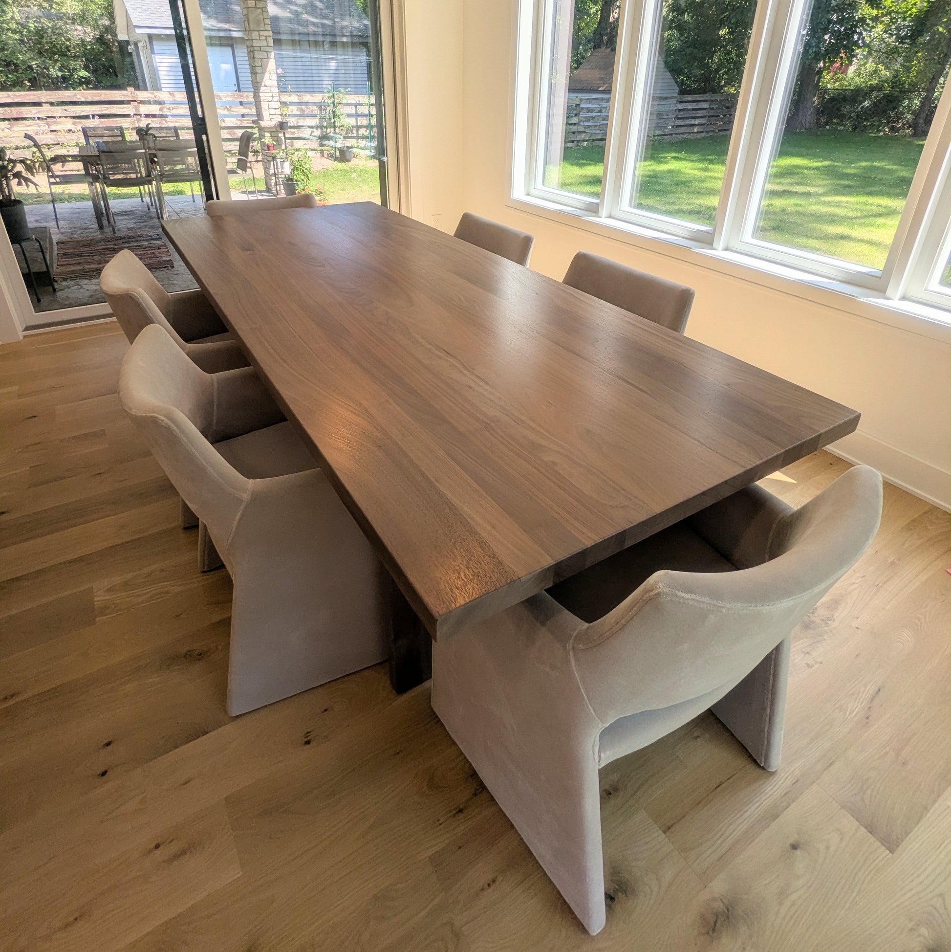A modern dining room featuring a long rectangular wooden table with six upholstered dining chairs in a light beige fabric, set in a bright room with large windows overlooking a green lawn.