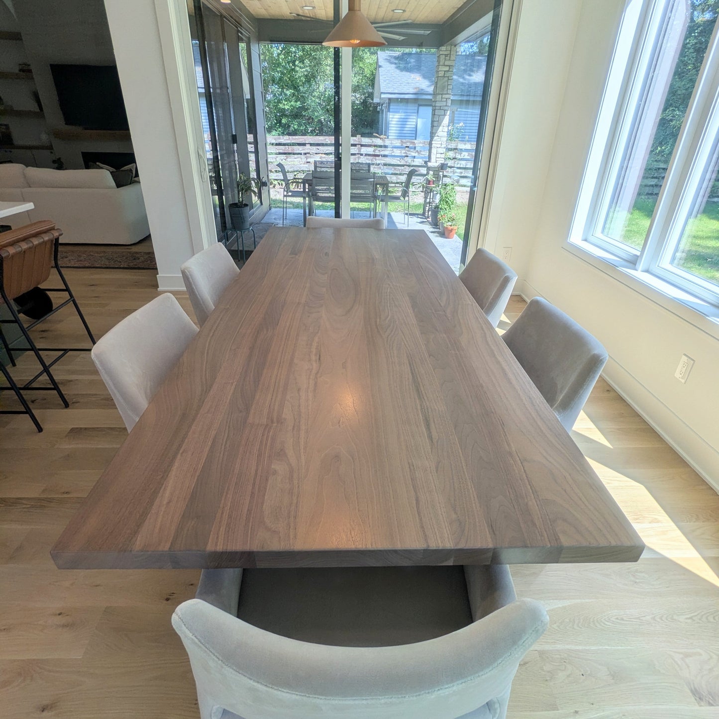 A long rectangular wooden dining table with a medium brown wood grain finish, surrounded by six light gray upholstered dining chairs in a bright, modern dining room with light wood floors and large windows.