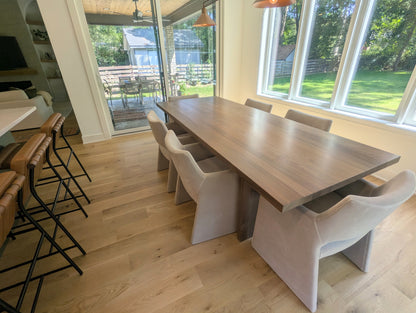 A modern dining room scene featuring a long rectangular wooden dining table surrounded by six upholstered dining chairs in a light beige fabric, set on light oak wood floors next to a kitchen island with brown leather bar stools.