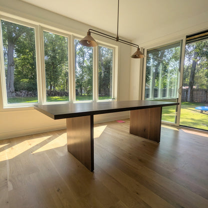 A modern rectangular dining table with a dark top and wood-toned block legs, placed in a bright room with large windows, hardwood floors, and a hanging industrial-style pendant light.