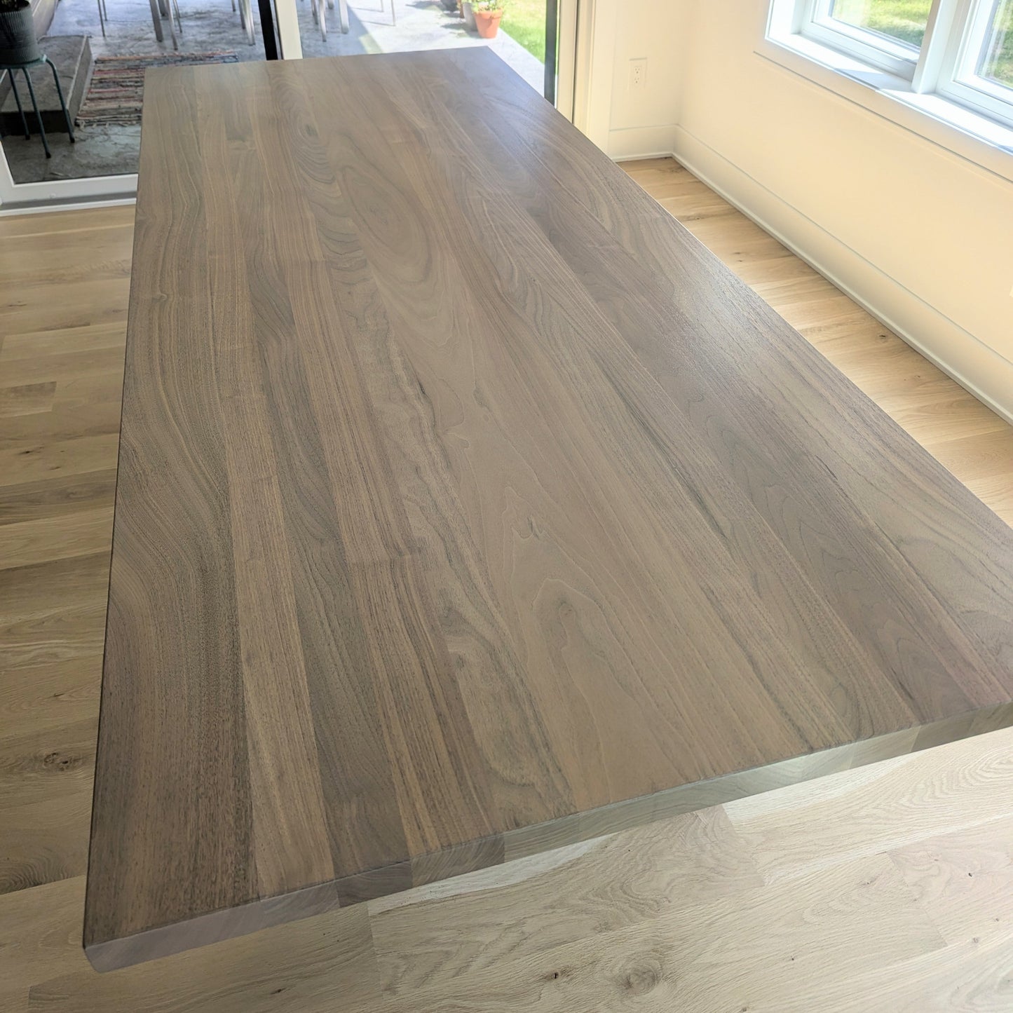 A large rectangular wooden dining table with a natural walnut-like grain, shown in a bright room with light wood floors and large windows looking out to a patio.