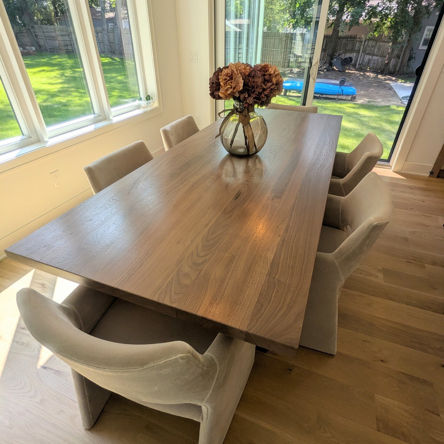 A rectangular wooden dining table with a natural oak finish, surrounded by six modern upholstered dining chairs in a light beige fabric, set in a bright room with hardwood floors and large windows overlooking a backyard.