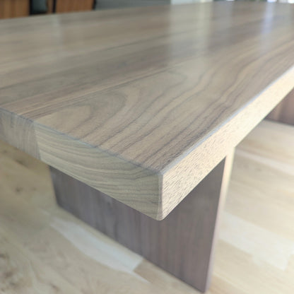 A close-up view of a modern wooden dining table with a thick, rounded rectangular top and a solid block base, shown in a bright, modern kitchen setting.